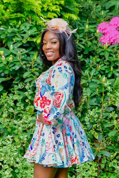 A woman in a colourful floral dress smiles against a backdrop of greenery and flowers.