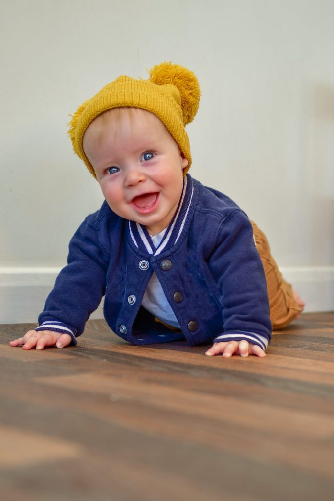 Baby with a yellow hat crawling on wooden floor, smiling at the camera.