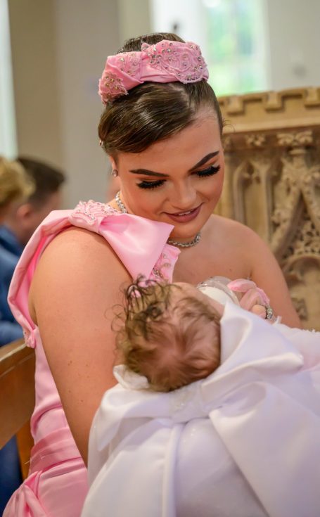 A mother in a pink dress smiles while holding her baby wrapped in a white Christening dress.