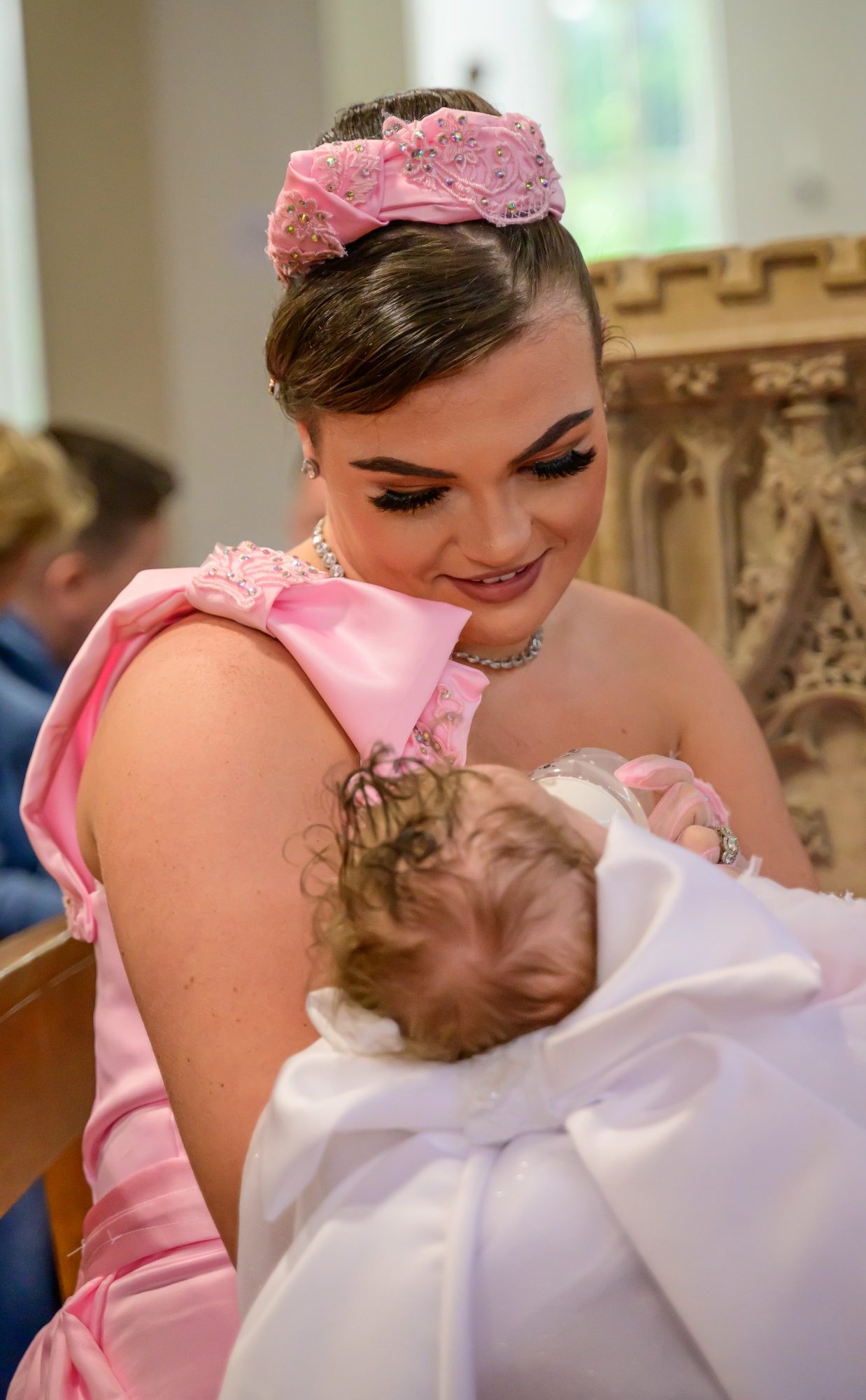 A mother in a pink dress smiles while holding her baby wrapped in a white Christening dress.