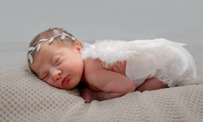 Newborn baby sleeping peacefully with a decorative headband and fluffy cape.