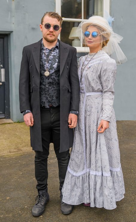 A man in sunglasses and a long coat stands beside a woman in a vintage dress and veil.